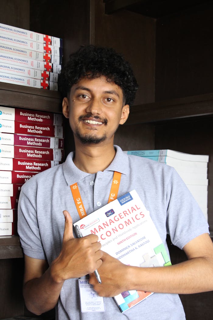 A student holding a textbook on Managerial Economics, smiling in a college library.