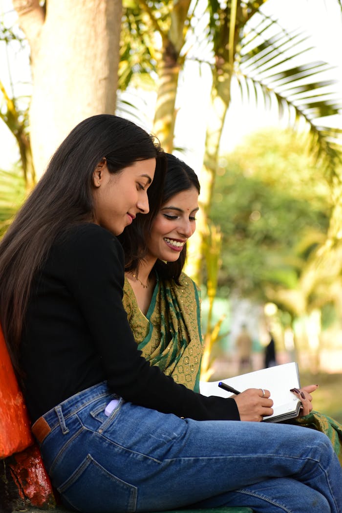 Two women smiling and writing in a notebook while sitting in a park with palm trees.